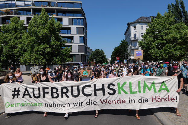 Eine Gruppe von Menschen mit Masken hält ein Transparent mit der Aufschrift "Aufbruchsklima" vor einem Gebäude mit Bäumen und einem klaren Himmel.