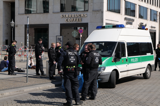 Polizeibeamte vor einem Starbucks-Café mit einem Van auf der rechten Seite und Menschen auf der linken Seite, mit einem Gebäude, einem Schild, einer Laterne und einem Zaun im Hintergrund.