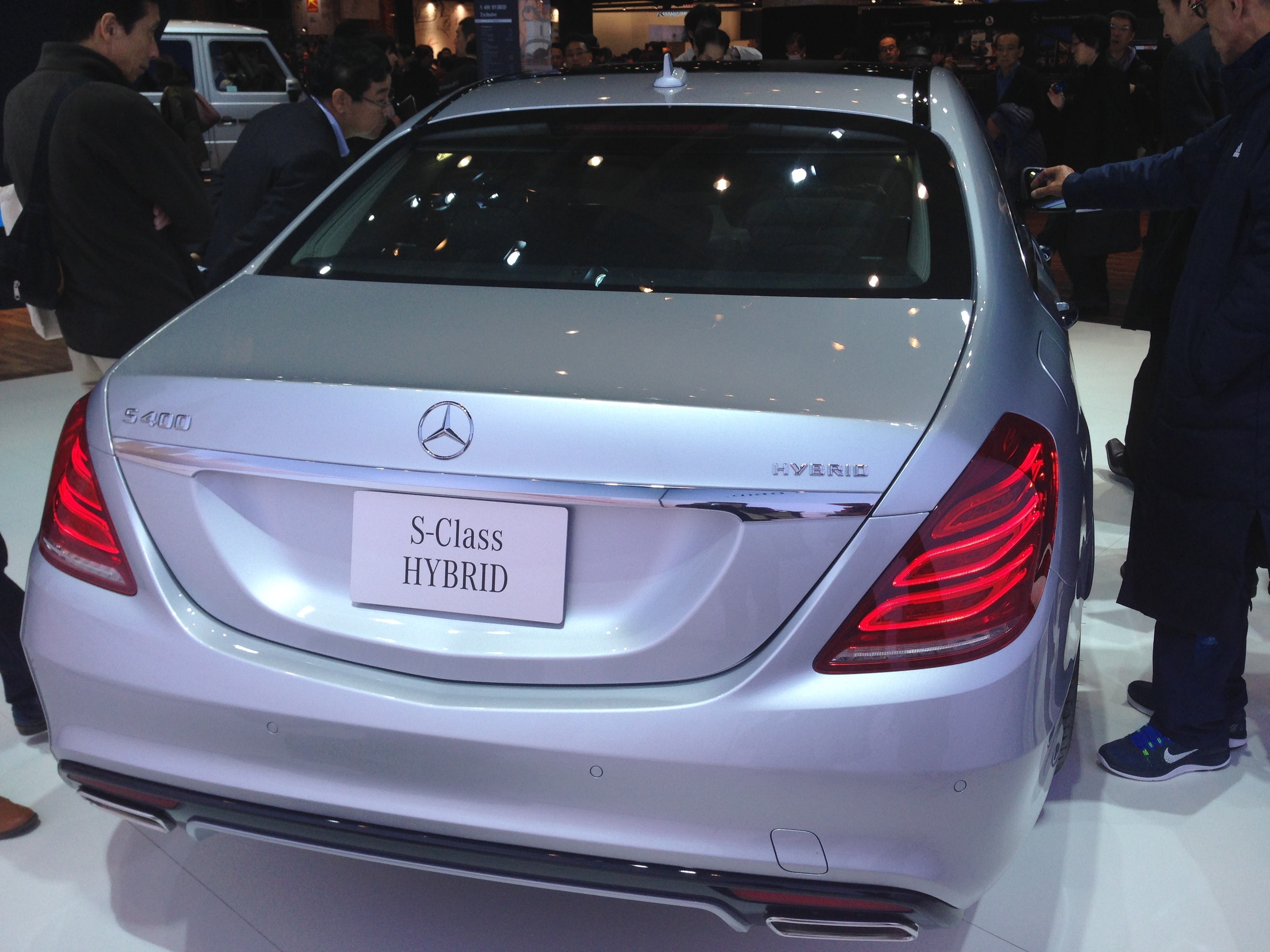 Silberner Mercedes S-Klasse Hybrid auf einer Automesse gezeigt, umgeben von Menschen in verschiedenenfarbigen Kleidern, mit anderen Fahrzeugen und Beleuchtung im Hintergrund.