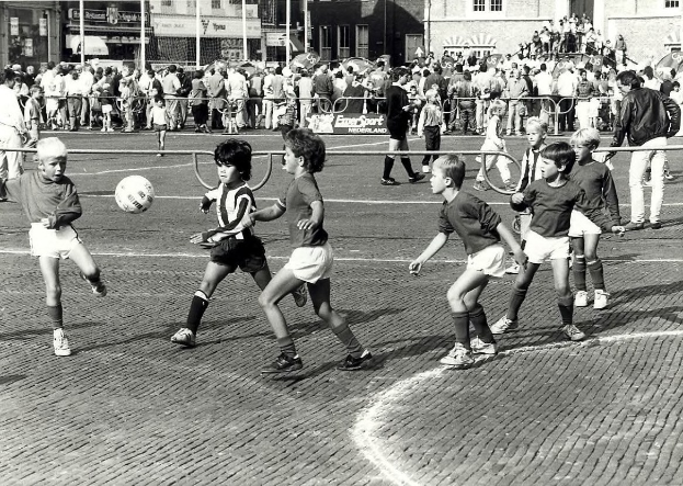 Schwarzes Bild von jungen Jungs, die Fussball auf der Straße spielen, umgeben von einer Zuschauermenge hinter einem Geländer, Gebäuden, Polen und Texttafeln im Hintergrund.
