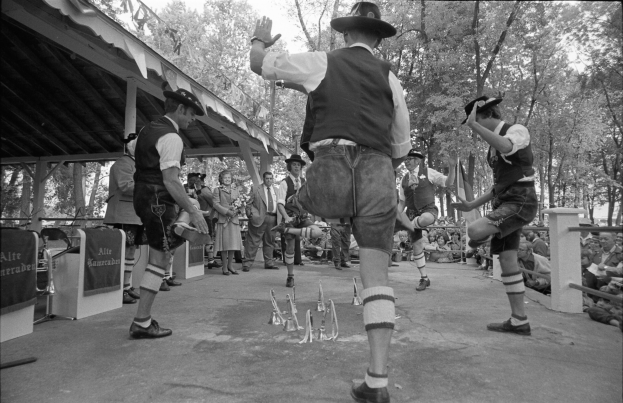 Eine Gruppe von Männern in traditioneller bayrischer Tracht tanzt auf einer Bühne vor einem sitzenden und stehenden Publikum, mit Bäumen und einer Hütte im Hintergrund, dargestellt in Schwarz-Weiß.