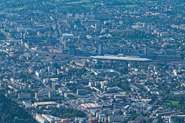 Eine Luftaufnahme einer belebten Stadt mit hohen, bunten Gebäuden, grünen Bäumen, gewundenen Straßen und saftig grünem Gras, mit Menschen, die spazieren gehen und Autos fahren.