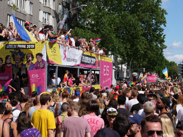 Eine große Menschenmenge marschiert während des Berlin Pride 2019 die Straße entlang, viele tragen Mützen und Schutzbrillen, einige halten Fahnen, mit Bannern, Laternenmasten, Bäumen und Gebäuden im Hintergrund unter einem bewölkten Himmel.