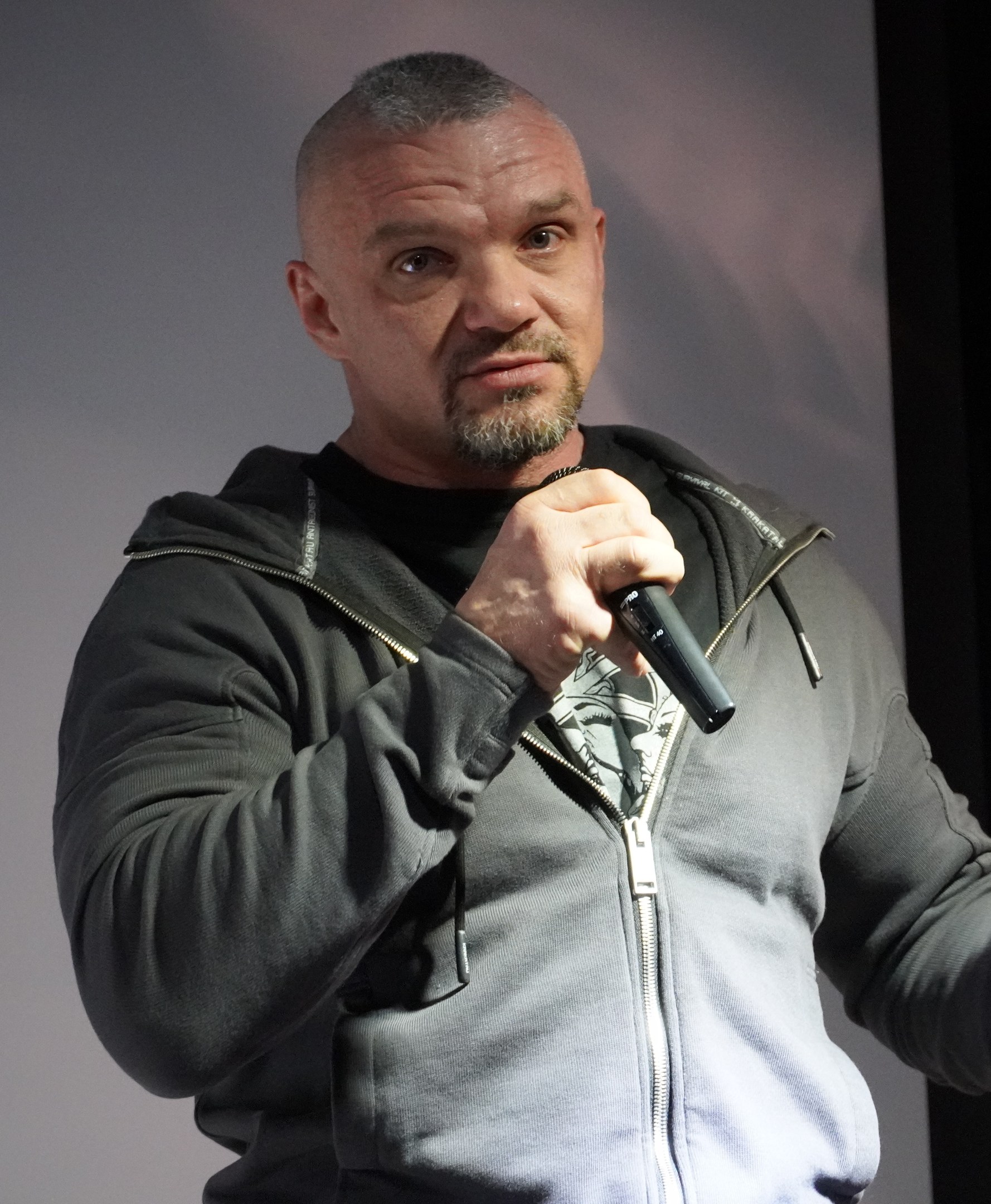 Ein Mann in einer schwarzen Kapuzenjacke und einem schwarzen Anzug mit einem weißen Hemd, glattem Haar und einem selbstbewussten Gesichtsausdruck hält ein Mikrofon, während er vor einer Wand steht.