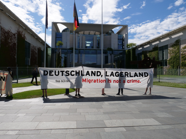 Gruppe von Menschen mit einem Transparent mit der Aufschrift "Deutschland, Lagerland Migration ist kein Verbrechen" vor einem Gebäude mit Fenstern, umgeben von Gras, einem Metallzaun, Pflanzen, Bäumen und Fahnen auf Stangen unter einem bewölkten Himmel.
