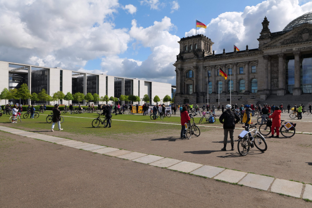 Eine Gruppe von Menschen, die Fahrräder vor dem Reichstaggebäude in Berlin, Deutschland, fahren. Das Gebäude ist mit Flaggen und Säulen geschmückt, der Boden ist grasbewachsen, es gibt Bäume im Hintergrund und ein bewölkter Himmel.