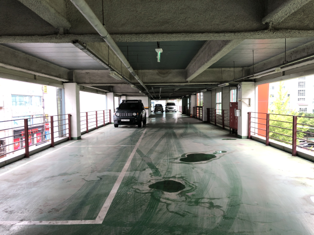 Ein mehrgeschossiges Parkhaus mit geparkten Autos, Geländern, Pfeilern, Deckenleuchten, freiliegenden Rohren und umliegenden Bäumen vor einem Hintergrund aus Gebäuden mit Fenstern in einer Stadtlandschaft.