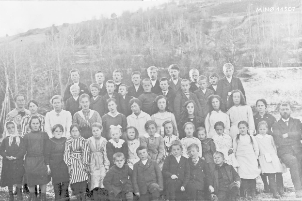 Ein Schwarz-Weiß-Foto einer Gruppe von Kindern, die vor Bäumen und einem Hügel posieren, mit der Beschriftung "1930-1940 Schulkinder" unten.