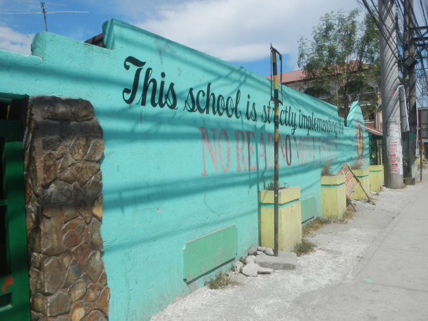 Grüne Wand mit Graffiti, das "Diese Schule ist keine Stadt" liest, vor einem bewölkten Himmel, umgeben von Gebäuden, Bäumen, Polen, Drähten und Brettern.