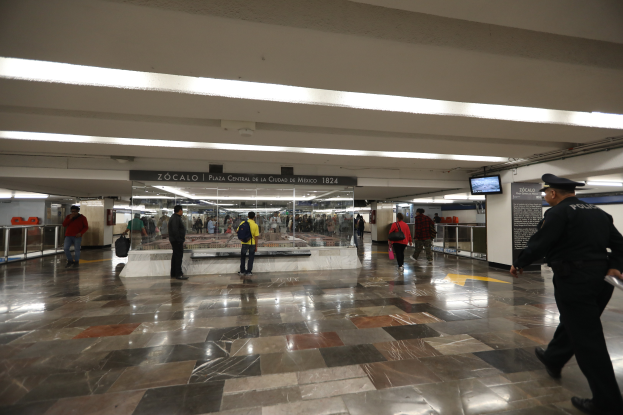 Eine Gruppe von Menschen durchquert eine belebte U-Bahn-Station, mit einem Polizeibeamten im Vordergrund, umgeben von Geländern, Bildschirmen und beleuchteten Schildern an der Decke.