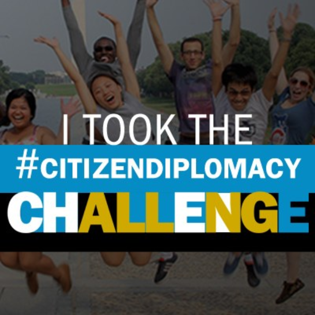 Eine Gruppe von Menschen, die in die Luft springen, mit dem Text "Ich habe die Staatsbürgerschaftsherausforderung bestanden" darüber, vor dem Hintergrund von Bäumen, Gebäuden und einem klaren blauen Himmel.