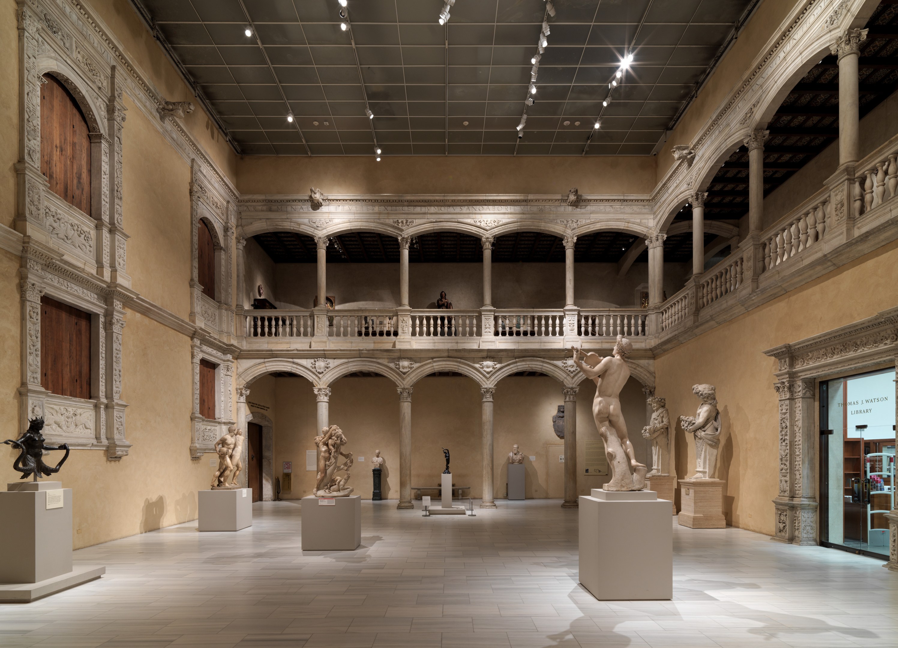 Großer Raum in einem Museum mit zahlreichen Statuen und Skulpturen auf Sockeln, architektonischen Elementen wie Wänden, Säulen und Geländern, Deckenleuchten und einigen Besuchern.