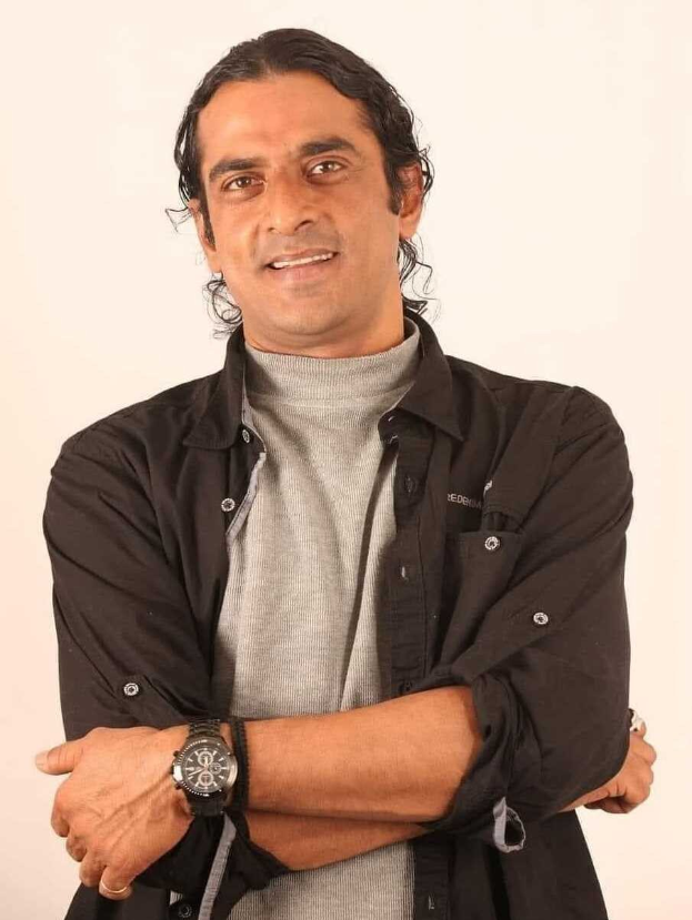 Ein Mann in einem schwarzen und aschfarbenen Kleid mit verschr├Ąnkten Armen, der eine Uhr tr├Ągt und gegen einen wei├čen Hintergrund steht und nachdenklich erscheint.