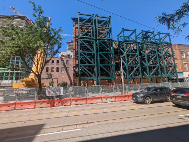 Großer Metallbrückenneubau an einer Baustelle mit geparkten Autos, Zaun, Bäumen, Gebäuden, einem Kran und Himmel im Hintergrund.