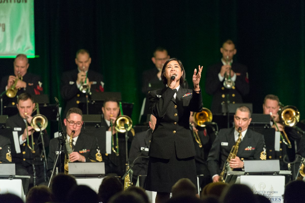 Eine Frau in Militäruniform singt in ein Mikrofon, während eine Band dahinter spielt, mit einem Lautsprecher und einem Banner im Hintergrund und einer Zuschauermenge darunter.