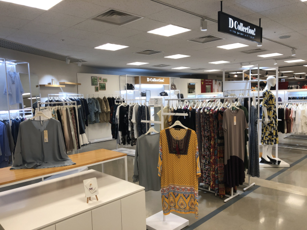 Innenansicht eines Kleiderladens mit Kleiderbügeln, Schaufensterpuppen und Tischen mit Artikeln, mit Wandtexttafeln und Deckenbeleuchtung, identifiziert als der neue D Collection Store im Mall of America.