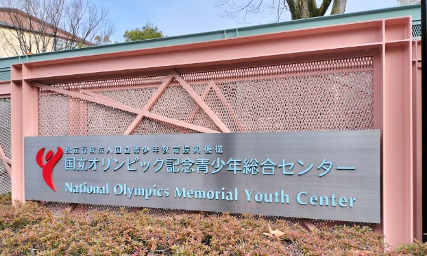 Außenansicht des Nationalen Olympischen Memorial Youth Center mit einem Namen auf der Wand, Grünzeug an der Basis, Bäumen und Gebäuden im Hintergrund und einem klaren blauen Himmel darüber.