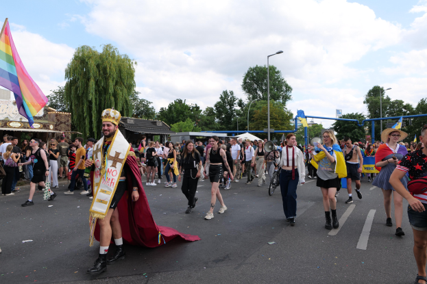 Eine Gruppe von Menschen marschiert bei der Gay Pride Parade 2018 mit einer Regenbogenflagge und Musikinstrumenten, während im Hintergrund Laternenpfähle, Bäume, Hütten und ein bewölkter Himmel zu sehen sind.