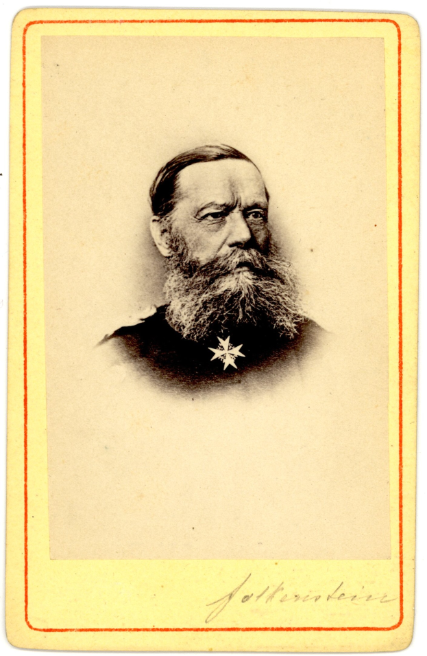 Altes Schwarz-Weiß-Foto eines bärtigen Mannes in Abendgarderobe mit einer Nadel an der Brust, der direkt in die Kamera schaut.