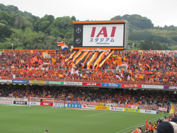 Ein Fußballspiel auf einem grünen Rasen mit einem Tor, umgeben von einer großen Zuschauermenge in den Stadionrängen, Fahnen, Schildern, einem großen Bildschirm, Bäumen und einem klaren blauen Himmel.