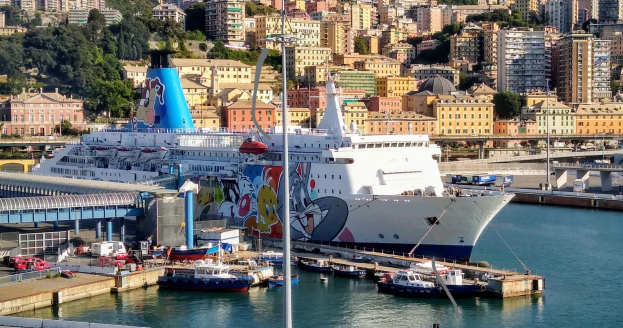 Großes Kreuzfahrtschiff an einem Stadtkai mit einer Brücke links, Fahrzeugen auf der Straße und Gebäuden und Bäumen im Hintergrund.
