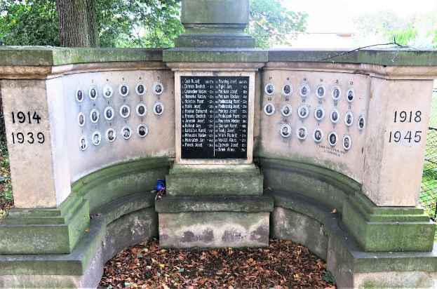 Ein Holocaust-Denkmal in einem jüdischen Friedhof in Berlin, das eine Wand mit Text und Zahlen zeigt, umgeben von Bäumen, einem Zaun und verstreuten trockenen Blättern.