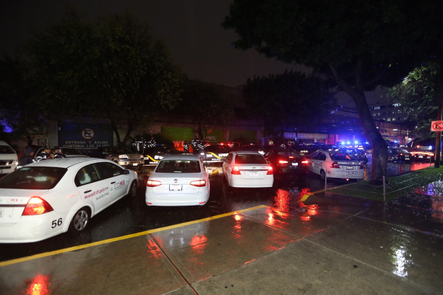 Eine Gruppe von Polizeiwagen auf einem Parkplatz bei Nacht mit einem Schild, Gras, Bäumen, Gebäuden, einer Fahne, Lichtern und einem bewölkten Himmel.