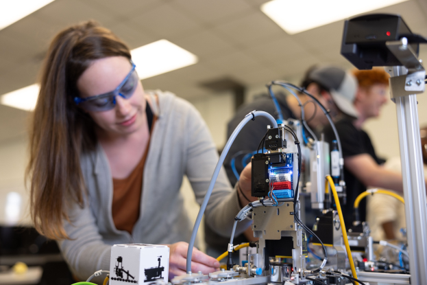 Eine Frau in Schutzbrille arbeitet an einem Roboter in einem hell erleuchteten Labor umgeben von Beobachtern, mit Werkzeugen und Komponenten auf dem Tisch.