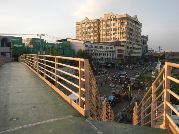 Eine Fußgängerbrücke mit Geländern überspannt eine vielbefahrene Straße mit Fahrzeugen und Fußgängern, mit Gebäuden, Bäumen, Strommasten mit Drähten und einem bewölkten Himmel im Hintergrund.