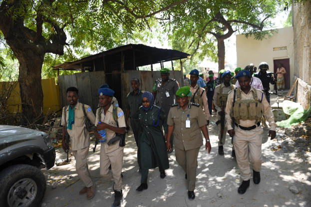 Eine Gruppe uniformierter Polizisten, einige bewaffnet, geht eine Straße entlang neben einem Auto, mit Bäumen, Schuppen, Wänden, Gebäuden und dem Himmel im Hintergrund.