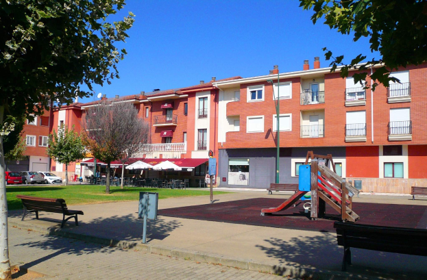 Ein Park mit einem Spielplatz, umgeben von Gebäuden, mit Bänken, Bäumen, Gras, einem Pfahl, einem Schild, einer Straßenlaterne, Fahrzeugen auf der Straße, Zelten, einer Gruppe von Menschen unter einem Dach und einer bewölkten Himmel.