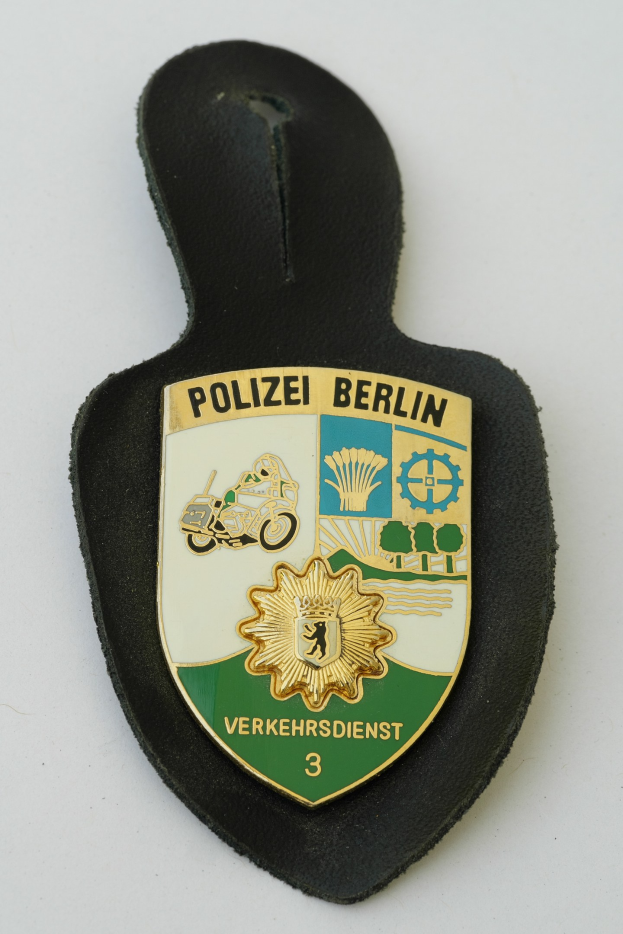 Ein schildförmiges Polizeibadged mit einem blauen Hintergrund, weißer Umrandung und einem zentralen weißen Stern, beschriftet mit 'Polizei Berlin' in schwarzer fetter Schrift, das auf einer Oberfläche ruht.