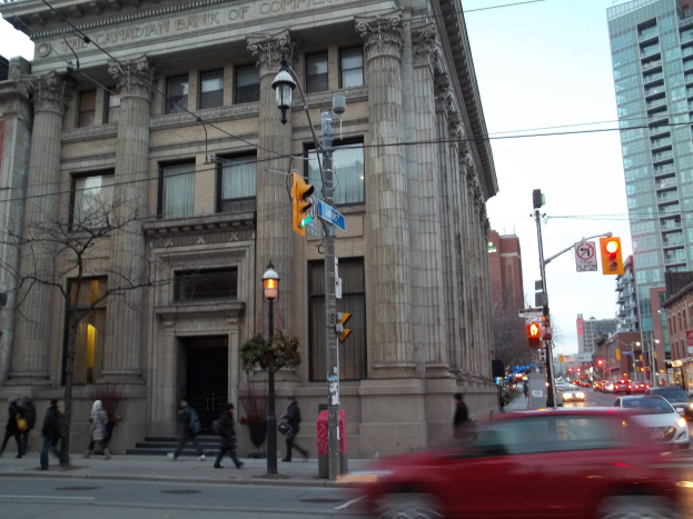 Eine belebte Stadtstraße mit Fahrzeugen, Fußgängern, Verkehrszeichen, Schildern, Bäumen und Gebäuden, mit einem roten Auto im Vordergrund und einem großen Gebäude links.