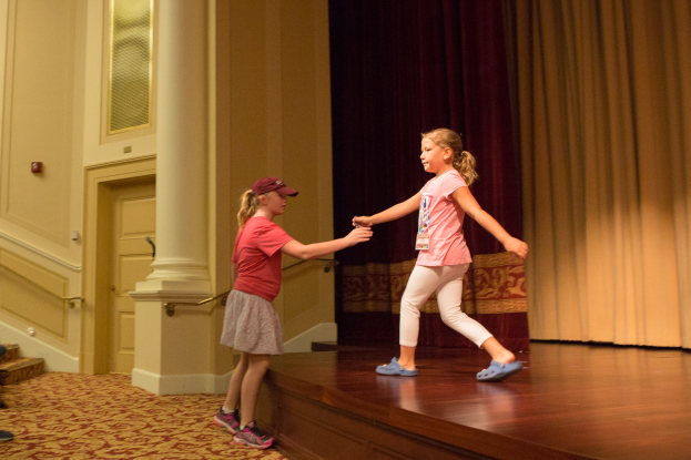 Zwei junge Mädchen tanzen auf einer Bühne vor einem Vorhang, mit einer Person, die auf einem Stuhl zu ihrer Linken sitzt, einer Säule und einer Wand im Hintergrund.
