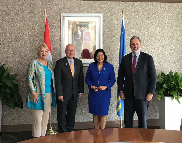 Eine Gruppe von Menschen, die fröhlich um einen Tisch mit Papieren stehen, umgeben von Fahnen, Topfpflanzen und einem Rahmenfoto an der Wand, was auf die Unterzeichnung eines U.S.-Canada Memorandum of Understanding hinweist.