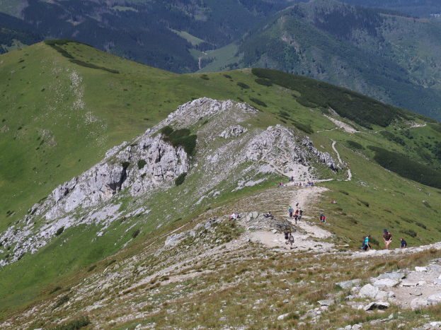 Menschen beim Wandern auf einem Berg mit grünem Gras und felsigem Gelände, Himmel im Hintergrund sichtbar.