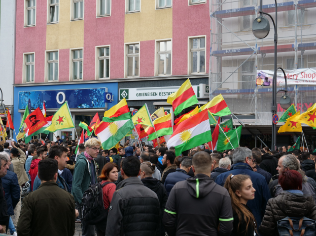 Eine große Gruppe von Menschen, die eine Straße entlanggehen und kurdische Flaggen schwenken, mit Laternenmasten und Gebäuden an der Straße und Texttafeln im Hintergrund.