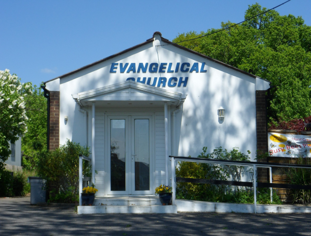 Evangelische Kirche mit Straße im Vordergrund, Grün im Hintergrund und einem klaren blauen Himmel darüber.