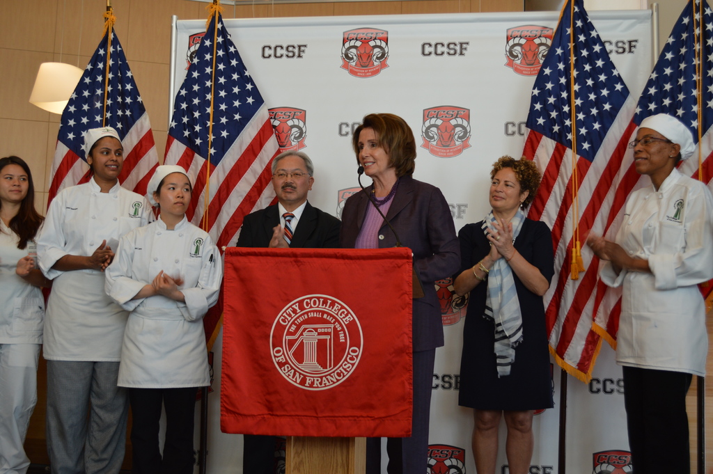 Eine Frau steht an einem Podium mit einem Mikrofon, lächelt, vor einem roten Tuch, mit Fahnen, einem Banner und einer Lampe dahinter und einer Wand im Hintergrund.