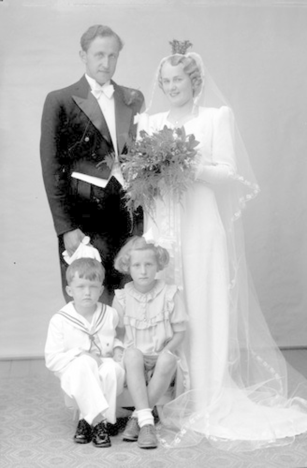 Schwarzes und weißes Foto einer Braut in einem weißen Kleid mit einem Bukett, Bräutigam neben ihr lächelnd und zwei Kinder vor ihnen sitzend.