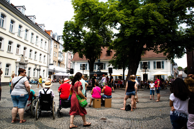Eine Gruppe von Menschen, darunter einige im Rollstuhl, geht eine Kopfsteinpflasterstraße in der Altstadt von Heidelberg entlang, mit Bäumen, Gebäuden, Laternen und einer Statue im Hintergrund unter einem bewölkten Himmel.