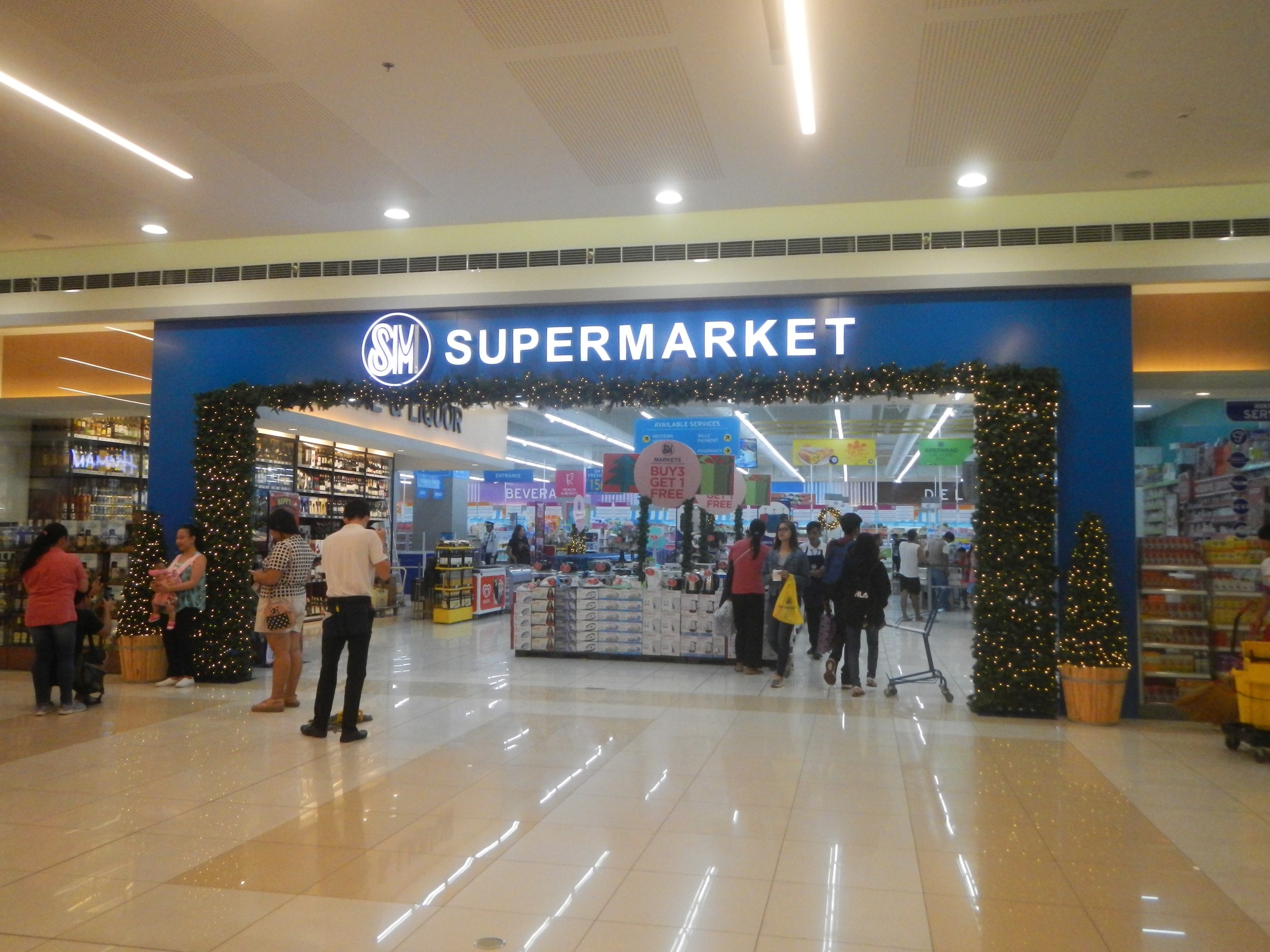 Gruppe von Menschen vor einem Supermarkt mit Einkaufstägern, einigen mit Einkaufstägern, mit einem Einkaufswagen, Topfpflanze, Regalen, Schildern und Deckenleuchten.