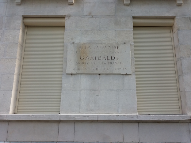 Gebäude mit zwei Fenstern und Läden, das eine Gedenktafel an der Seite trägt.