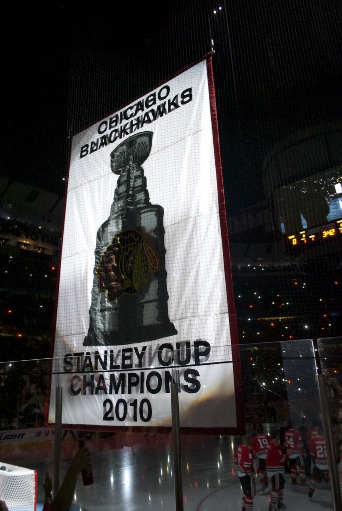 Eine nächtliche Stadion-Szene mit einem großen Banner in der Mitte, Zuschauern im Hintergrund, einem Glas auf dem Boden und einigen Spielern auf dem Feld, beleuchtet von Stadionlampen.