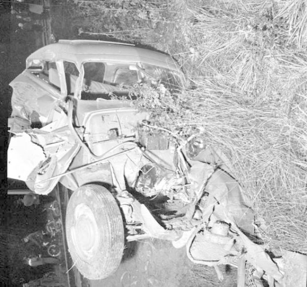 Schwarze und weiße Fotografie eines stark beschädigten Autos mit eingedellter Front und zersplitterten Fenstern am Straßenrand, mit einem Baum und verstreuten Gegenständen im Hintergrund.