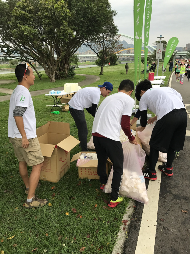 Group of people on a roadside during a charity run, surrounded by litter and banners, with trees, buildings, and a clear sky in the background.
