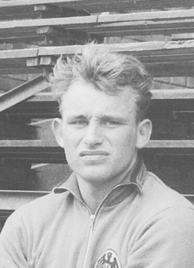 Schwarzes und weißes Foto eines Mannes in einer Jacke vor gestapelten Holzplanken, der angeblich der legendäre deutsche Füßballer ist.