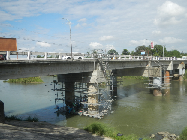 Brücke mit Gerüst über einem Fluss, Fahrzeuge vorhanden, umgeben von Gras, Felsen, Bäumen und einem bewölkten Himmel.