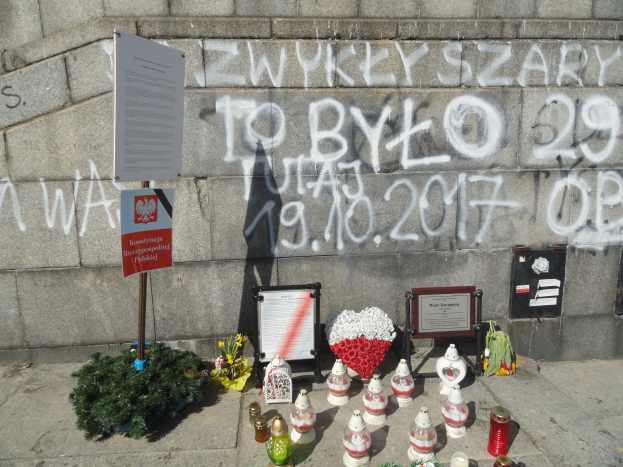 Gedenkstätte mit Blumen, Kerzen und Zetteln vor einer mit Graffiti bedeckten Steinwand, wahrscheinlich zur Erinnerung an Holocaust-Opfer.