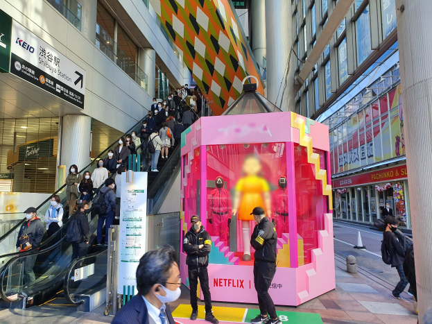 Eine Gruppe von Menschen steht vor einem Gebäude mit Glaswänden, Säulen und Texttafeln, einige tragen Masken, während hinter ihnen eine Rolltreppe mit Menschen nach oben fährt; eine pinkfarbene Box in der Mitte zeigt Schaufensterpuppen und trägt den Text "Netflix's new Netflix booth in Tokyo."